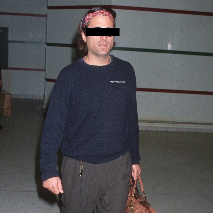 A man wearing a navy blue crewneck sweatshirt with 'ZEBRAKIDS' printed in white on the chest, paired with dark gray sweatpants and a red patterned headband. He is holding a brown leather bag and standing in an indoor hallway.