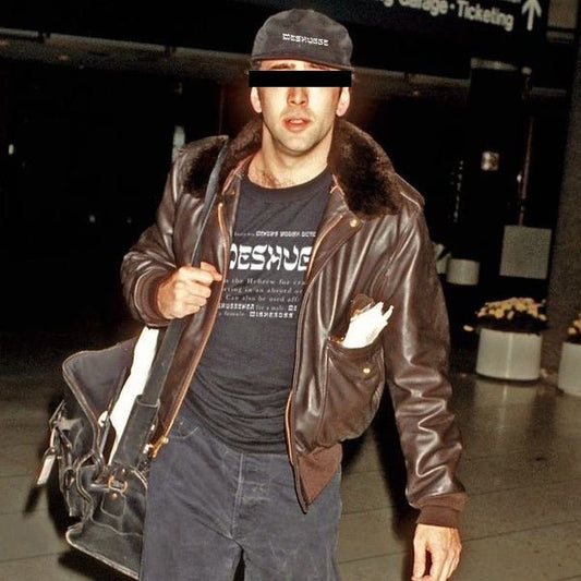 A man wearing a dark brown leather aviator-style jacket with a dark fur collar, a black t-shirt, and blue jeans, carrying a black shoulder bag in an airport setting.