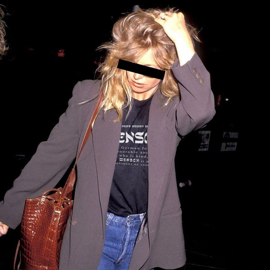 A woman in a dark gray blazer, black graphic t-shirt, and blue jeans carrying a brown crocodile-embossed shoulder bag, photographed at night with her face obscured by a black bar.
