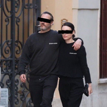 A man and a woman walking arm in arm wearing matching black crewneck sweatshirts with the brand name 'Meshugge' printed in white on the chest, set against an outdoor urban background.
