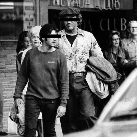 Two men walking outdoors with a building and people in the background