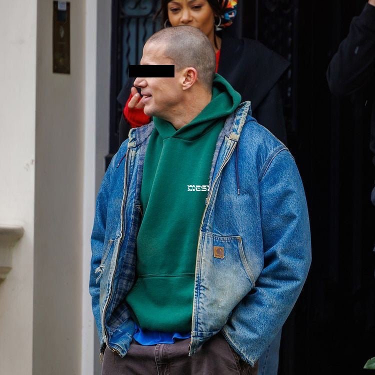 A man wearing a green hoodie under a blue denim jacket, standing outdoors in front of a building entrance. The face is blurred, and another person is partially visible behind him.