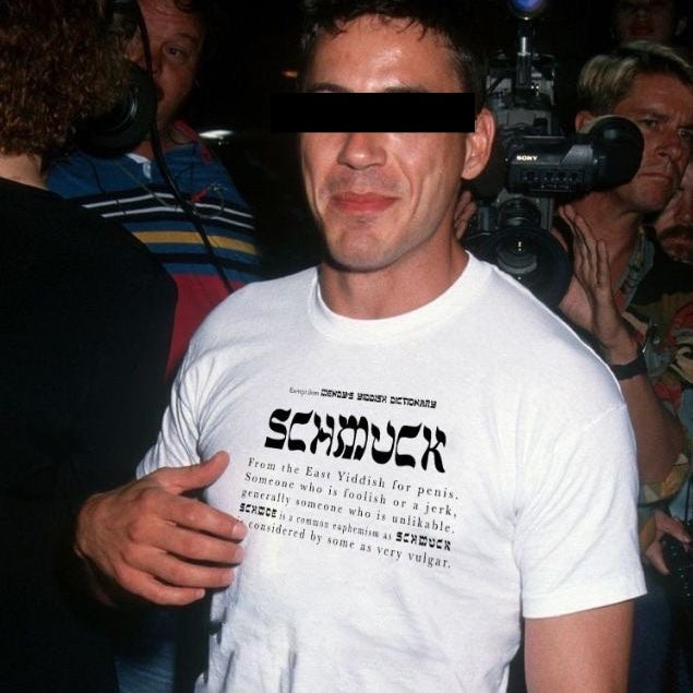A man wearing a white graphic t-shirt with black text that reads 'SCHMUCK' and a dictionary-style definition, photographed in a crowded indoor setting with a camera and other people visible.