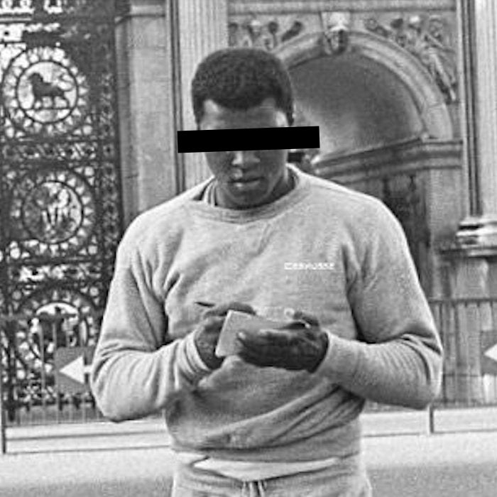 A black and white photo of a man in a sweatshirt standing outdoors in front of an ornate gate and building archway, holding a small card or object in both hands. His face is obscured by a black bar.