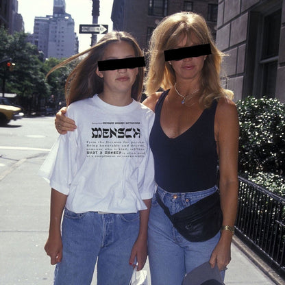Two women standing on a city sidewalk, with the younger woman wearing a white graphic T-shirt and blue jeans. The T-shirt features the word 'MENSCH' in large black letters with smaller text underneath.