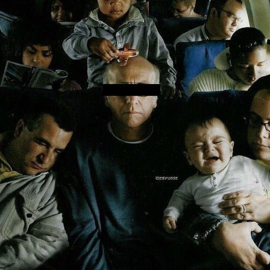 A dark, crowded scene showing several people in what appears to be an airplane or train setting. A man in the center wears a black eye mask, with a child above him holding a cup and a baby on the right being held by an adult.