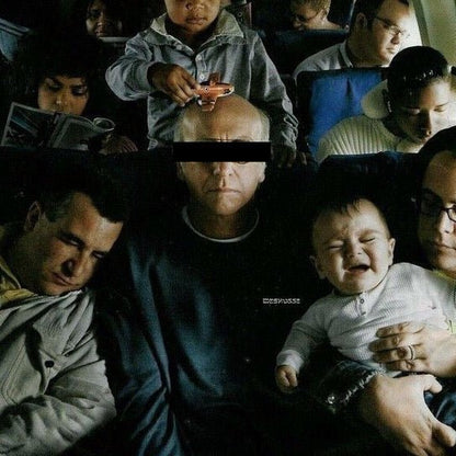 A dark, crowded scene showing several people in what appears to be an airplane or train setting. A man in the center wears a black eye mask, with a child above him holding a cup and a baby on the right being held by an adult.