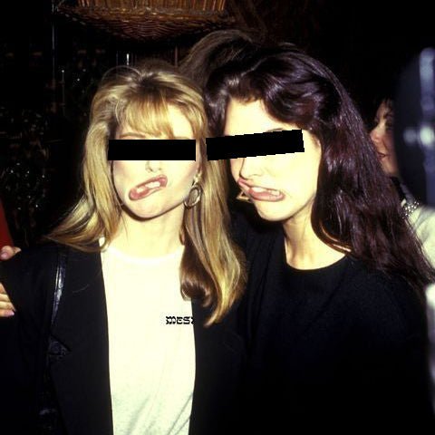 Two women posing closely together in a dark indoor setting, with their eyes obscured by black bars. One wears a black blazer over a white top with a small black logo, and the other wears a black top.
