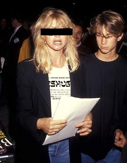 A blonde woman in a black blazer over a white graphic T-shirt and blue jeans holds papers, standing next to a young man in a black long-sleeve shirt at a social event. Her face is obscured by a black bar.