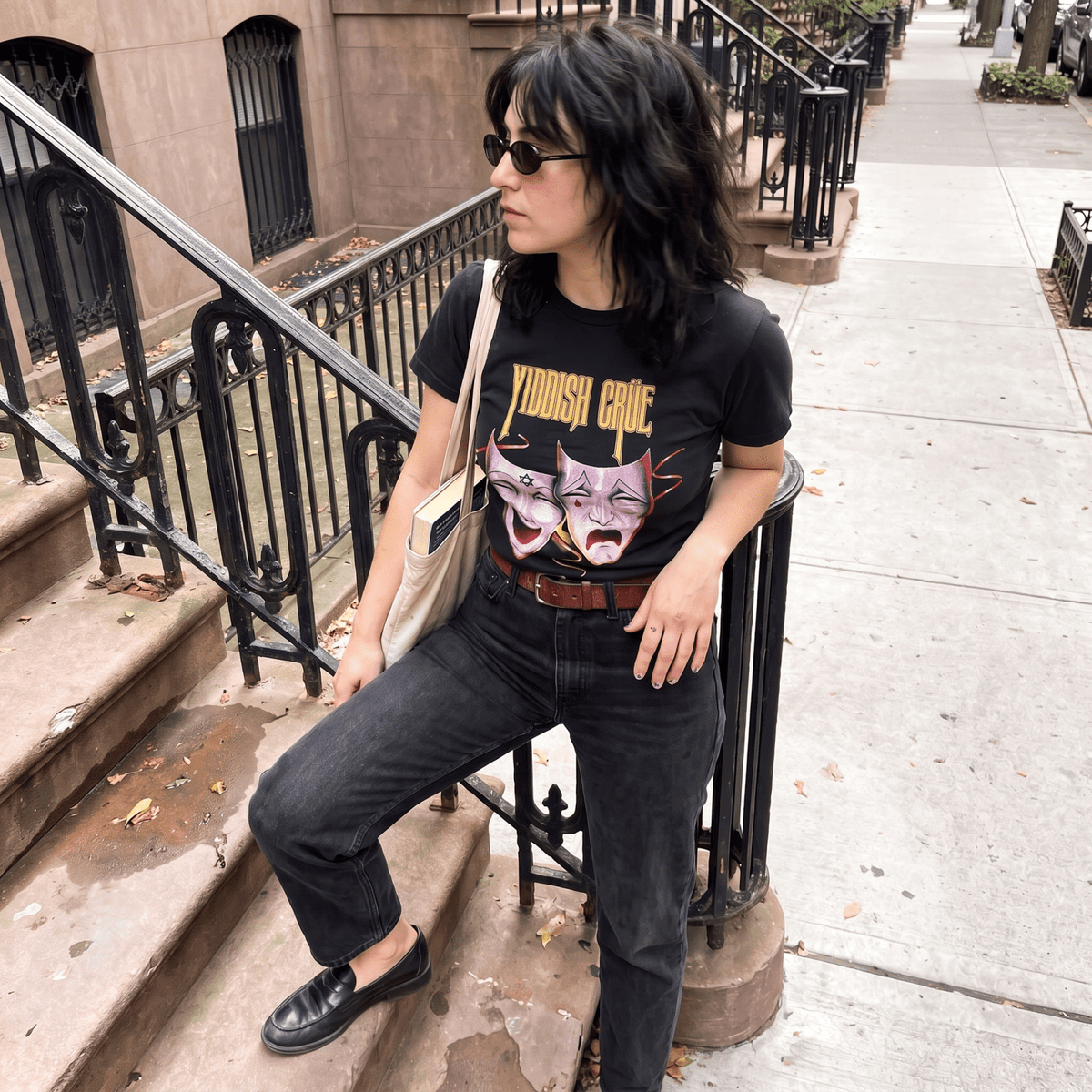 A woman wearing a black graphic t-shirt with 'YIDDISH CRUE' and a purple cat illustration, paired with black jeans and sunglasses, sitting on a city sidewalk next to a brownstone building.