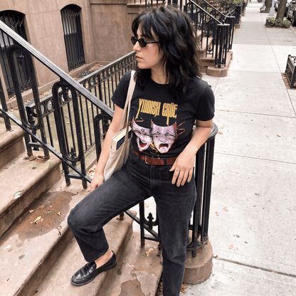 A woman wearing a black graphic t-shirt with 'YIDDISH CRUE' and a purple cat illustration, paired with black jeans and sunglasses, sitting on a city sidewalk next to a brownstone building.