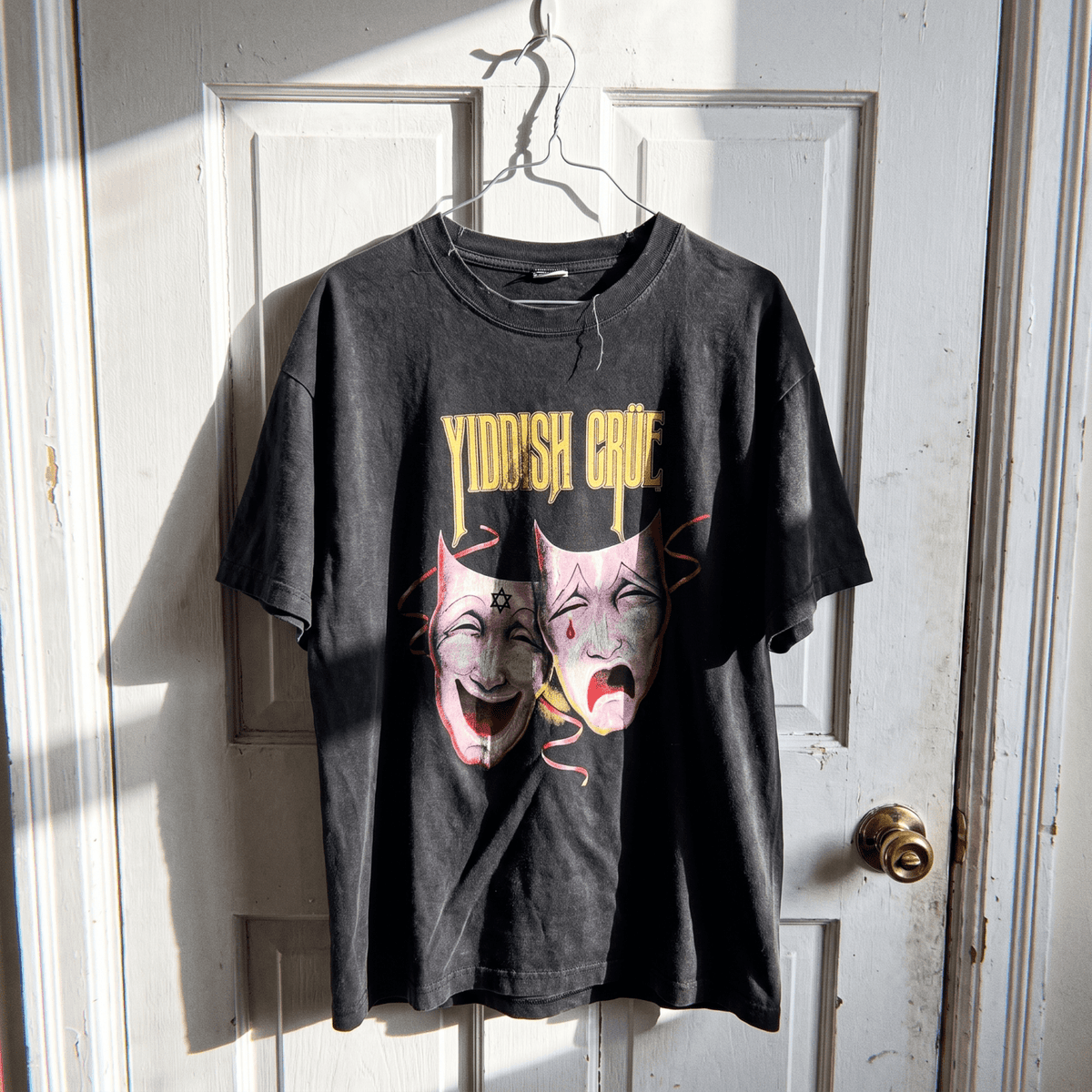 A black washed graphic t-shirt hanging on a metal hanger against a white paneled door. The graphic features the text 'YIDDISH CRUE' in yellow above two theatrical comedy and tragedy masks in pink and yellow tones.