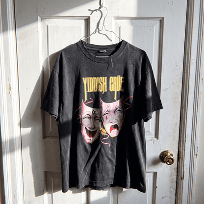 A black washed graphic t-shirt hanging on a metal hanger against a white paneled door. The graphic features the text 'YIDDISH CRUE' in yellow above two theatrical comedy and tragedy masks in pink and yellow tones.
