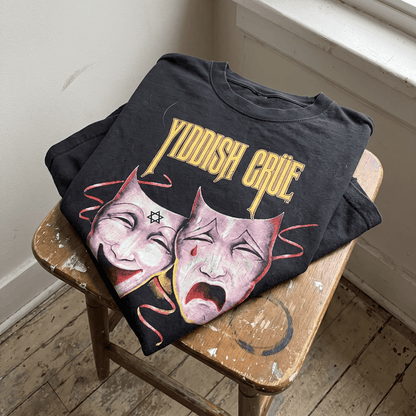 A black graphic t-shirt with the words 'YIDDISH CRUE' in yellow and an illustration of two theatrical masks, resting on a worn wooden stool in a room corner.
