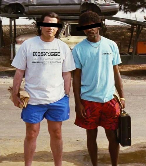 Two men standing outdoors in a dusty, rustic setting. The man on the left wears a white t-shirt with a black graphic print and blue shorts, holding a brown paper bag.