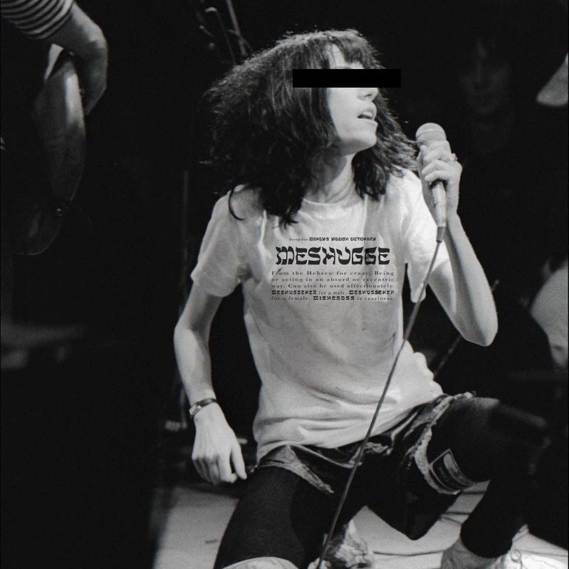 A black and white photo of a person on a stage holding a microphone, wearing a white graphic t-shirt with the word 'Moshpit' printed on the front, ripped jeans, and dark leggings.