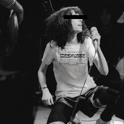 A black and white photo of a person on a stage holding a microphone, wearing a white graphic t-shirt with the word 'Moshpit' printed on the front, ripped jeans, and dark leggings.