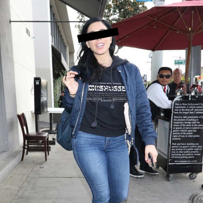 A woman walking on a city sidewalk wearing a black graphic T-shirt under a navy blue hoodie and blue jeans. The T-shirt has white text reading 'Meshugge' with smaller text below.